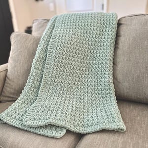 May include: A soft, light teal blanket with a textured, raised pattern is draped over a gray sofa. The blanket's color fades from a deeper teal to a lighter shade, creating a subtle ombre effect. The blanket appears cozy and inviting.