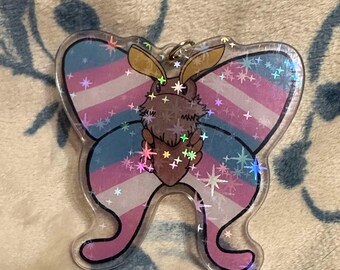 May include: A clear acrylic charm featuring a brown and tan butterfly with a pink, blue and white striped pattern. The butterfly has a glittery finish and is surrounded by small white stars.