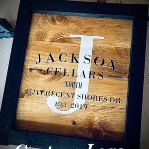 May include: Wooden sign in a black frame. The sign reads "JACKSON CELLARS NORTH 2524 CRECENT SHORES DR Est. 2019" with a white "J". The words "Custom Logo" are at the bottom of the sign.