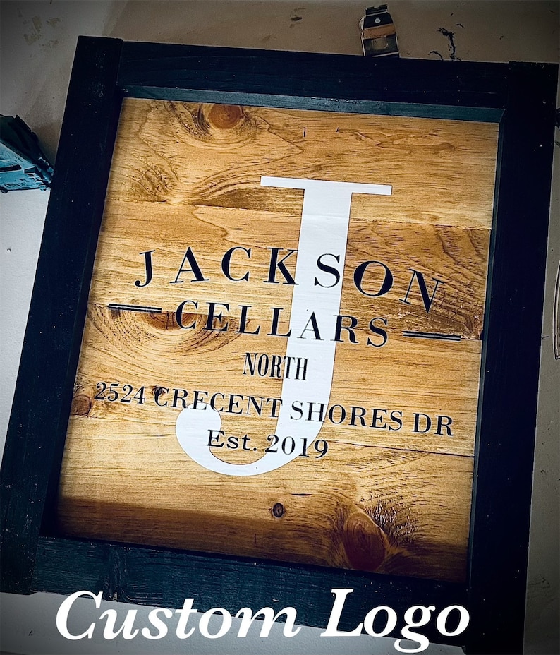 May include: Wooden sign in a black frame. The sign reads "JACKSON CELLARS NORTH 2524 CRECENT SHORES DR Est. 2019" with a white "J". The words "Custom Logo" are at the bottom of the sign.