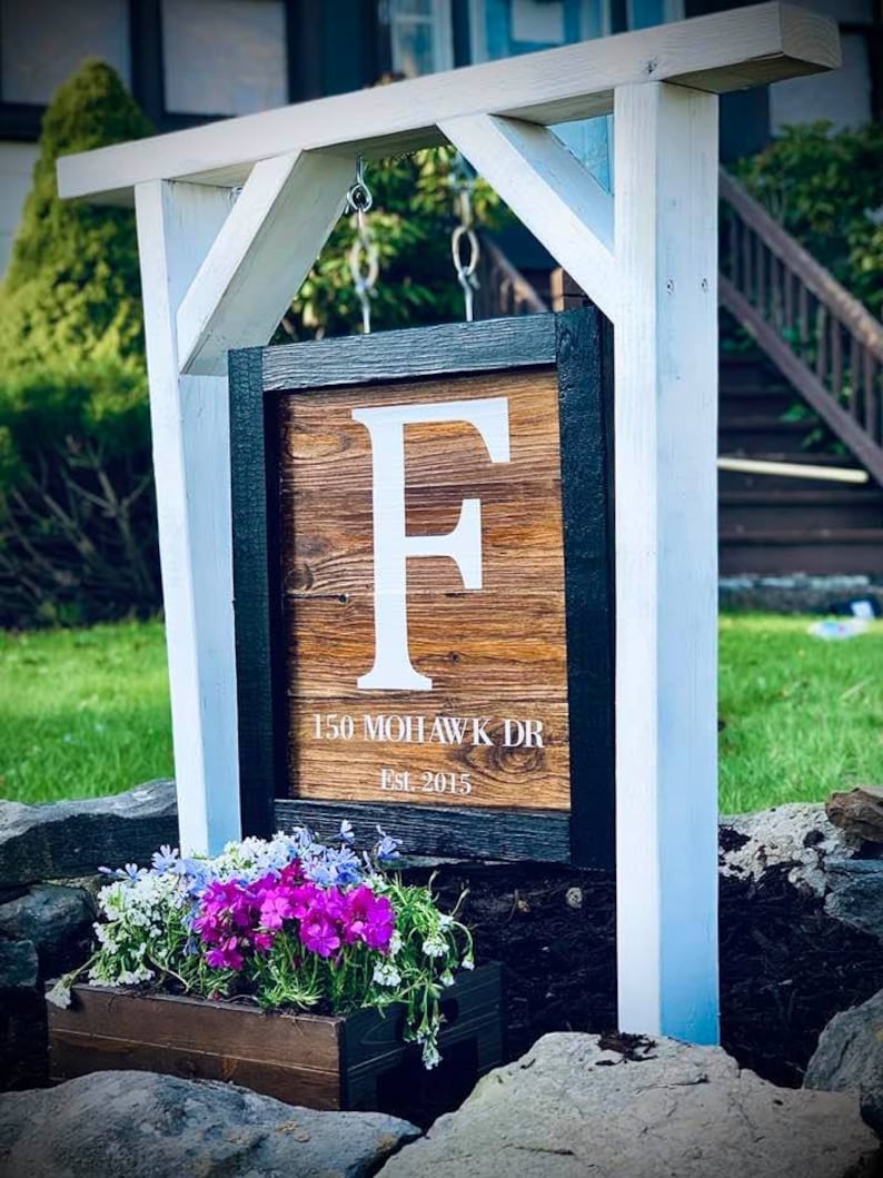 May include: A wooden address sign featuring the letter "F" in white, framed in black, and mounted on a white post. The sign reads "150 MOHAWK DR" and "Est. 2015". A wooden planter box with colourful flowers is positioned below the sign.
