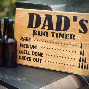 May include: A wooden sign with the text "Dad's BBQ Timer" and a list of doneness levels for grilling meat, including "Rare", "Medium", "Well Done", and "Order Out". The sign is decorated with small bottle silhouettes.