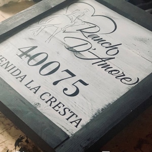 May include: Square wooden sign with a black frame and a white-washed background. The sign features a horse and rider graphic, the words "Ranch D'Amore", the number "40075", and the text "Avenida La Cresta". The words "Custom Logo" are at the bottom.