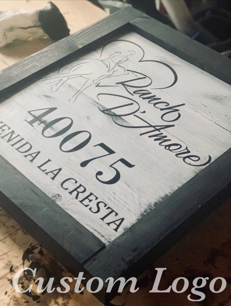 May include: Square wooden sign with a black frame and a white-washed background. The sign features a horse and rider graphic, the words "Ranch D'Amore", the number "40075", and the text "Avenida La Cresta". The words "Custom Logo" are at the bottom.