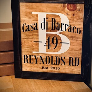 May include: Wooden framed sign with the text "Casa di Barraco 49 Reynolds Rd Est. 2010" in black and white. The sign has a large white letter "B" in the center. The frame is dark brown.