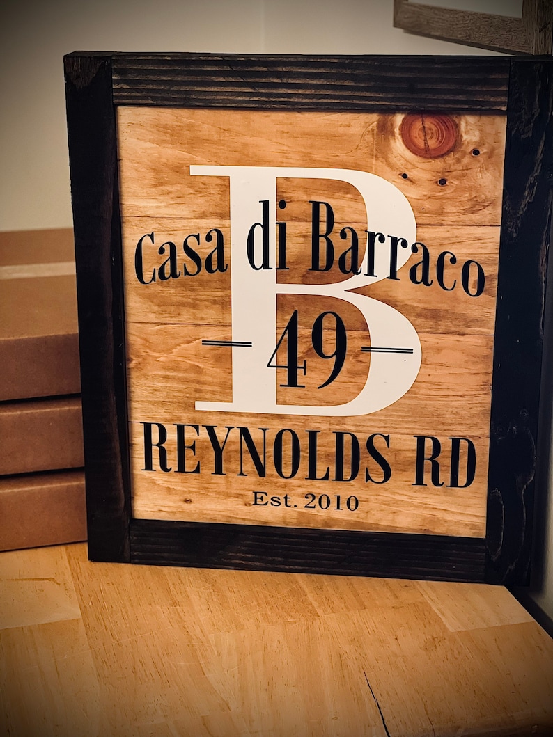 May include: Wooden framed sign with the text "Casa di Barraco 49 Reynolds Rd Est. 2010" in black and white. The sign has a large white letter "B" in the center. The frame is dark brown.