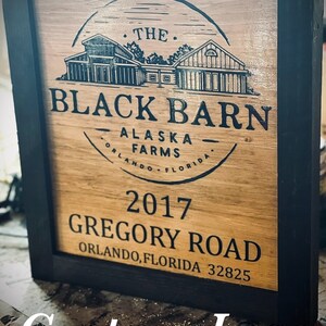 May include: Wooden sign in a black frame. The sign displays the text "THE BLACK BARN ALASKA FARMS ORLANDO FLORIDA" and "2017 GREGORY ROAD ORLANDO, FLORIDA 32825". It also includes a custom logo.