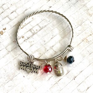 May include: Silver bangle bracelet with charms, including a baseball charm with the words "Baseball Mom" and a heart, a red crystal charm, and a silver baseball cap charm.