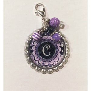 May include: A silver-colored bottle cap charm with a decorative purple and black design, featuring the letter "C". The charm is adorned with purple and white striped beads, purple beads, and a silver clasp.