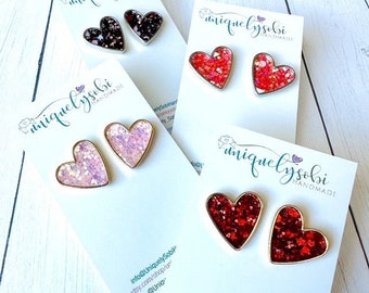 May include: Four pairs of heart-shaped earrings with glitter embedded in the resin. The earrings are in four different colors: pink, red, black, and a mix of red and pink. The earrings are on white cards with the text "uniquelylysobi handmade" printed on them.