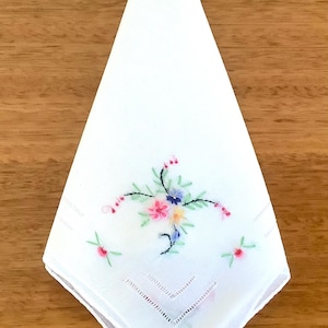 May include: White handkerchief with a floral embroidery design. The embroidery features pink, blue, and yellow flowers with green leaves and stems. The handkerchief is folded in a triangle shape.