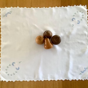 May include: A white rectangular tablecloth with embroidered blue and white floral designs in each corner and a delicate lace trim. Four wooden decorative objects in shades of brown are arranged on the tablecloth.