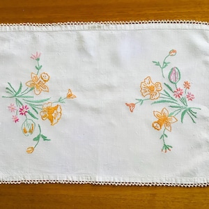 May include: White rectangular embroidered placemat with a crocheted edge. The placemat features floral embroidery in orange, pink, and green. The flowers are arranged in two clusters on opposite sides of the placemat.