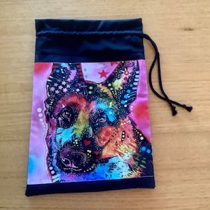 May include: A black drawstring bag with a colorful graphic of a German Shepherd dog on the front. The dog is depicted in a vibrant, abstract style with pink, blue, yellow, and green colors.