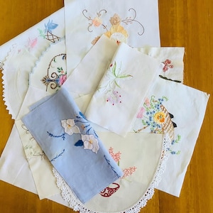 May include: A selection of vintage embroidered linens, including handkerchiefs and doilies. The textiles display floral patterns in shades of pink, blue, and yellow. Some pieces have lace edging and detailed cross-stitch work.
