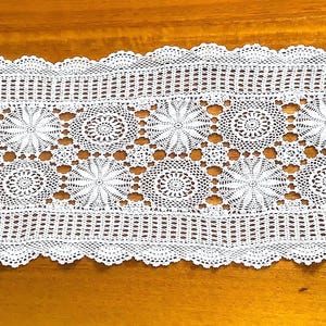 May include: White, rectangular crochet table runner with a scalloped edge. The center features a repeating pattern of floral and circular designs. The openwork design allows the surface beneath to show through. A decorative piece for a dining table or other surface.