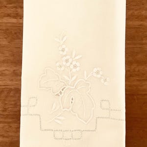 Peut inclure: Une serviette de toilette en lin blanc avec une broderie blanche délicate représentant un motif floral. La broderie présente une tige feuillue avec de petites fleurs et une feuille plus grande.