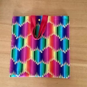 May include: A reusable tote bag with red handles and a colourful pattern of pencils. The pencils are arranged in a rainbow pattern and have a light brown wood finish.