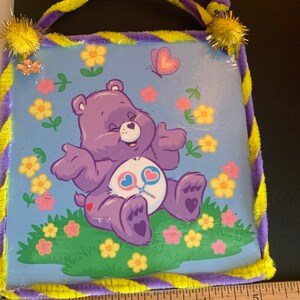 May include: A purple Care Bear with a pink heart on its chest sits on a bed of green grass with yellow and pink flowers. The bear is holding two pink and blue lollipop sticks. A butterfly with pink wings flies above the bear. The image is framed with a yellow and purple striped border.