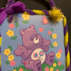 May include: A purple Care Bear with a heart on its chest and a lollipop in its hand sits on a patch of green grass with yellow and pink flowers. The Care Bear is smiling and has its arms outstretched. The background is a light blue sky with a butterfly flying above the Care Bear.