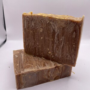May include: Two rectangular bars of handmade soap. The soap is brown with lighter, swirled patterns. The top edges have a golden hue. The bars are stacked, highlighting the texture and colour variations. Each bar is approximately 10 cm long.