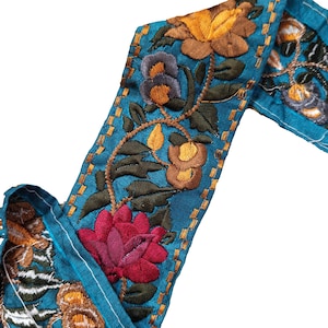 Pode incluir: Tecido azul-turquesa com bordado floral intrincado. O design apresenta uma variedade de flores em tons de vermelho, amarelo, castanho e roxo, com folhas e caules verdes. As bordas são costuradas com fio dourado, criando uma borda decorativa.