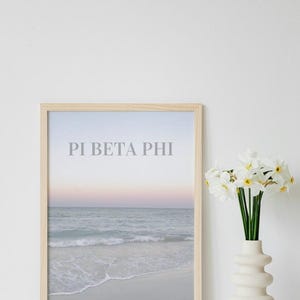 May include: A framed print featuring a light blue and white beach scene with the text "Pi Beta Phi" in grey. The print is displayed on a white surface with a white vase of white daffodils.