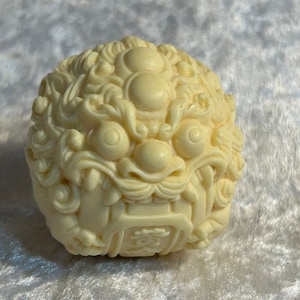 May include: A white carved sphere featuring an intricate design of a lion's face. The sphere is crafted from a smooth, ivory-like material.