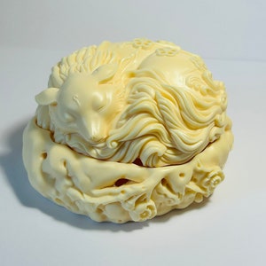 May include: Ivory-coloured decorative box with a sleeping fox design. The box features intricate carvings of the fox, floral patterns, and flowing details. The fox is curled up on top of the box, which is round and has a lid.