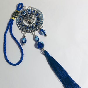 May include: A silver charm featuring an owl design with blue evil eye beads hanging from a blue tassel. The charm is attached to a blue cord.