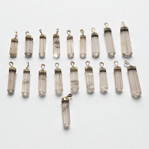 Może przedstawiać: Kolekcja wisiorków z przezroczystego kryształu kwarcu ze złotymi metalowymi nakładkami i pętelkami. Cylindryczne wisiorki różnią się nieznacznie rozmiarem i przejrzystością. Tło jest jednolicie białe.