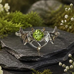Puede incluir: Un anillo de tono plateado con una piedra preciosa redonda de color verde lima en el centro, flanqueada por dos alas de murciélago detalladas. El diseño del anillo incorpora un elemento de cola puntiaguda bajo la piedra, creando un motivo.