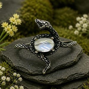 May include: A silver-toned ring featuring a coiled snake design with a textured, dark-oxidised finish. The snake's body wraps around a central, smooth, oval-shaped iridescent moonstone cabochon, creating a detailed and intricate metal band.