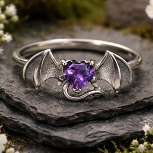 Puede incluir: Un anillo de plata con una piedra preciosa morada en forma de corazón en el centro, flanqueada por dos alas de murciélago detalladas. La banda es fina y metálica, con las alas extendiéndose desde la piedra morada facetada.