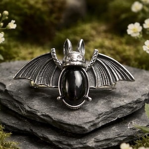 May include: A silver-toned metal brooch shaped like a bat with outstretched wings. The centre of the bat features a smooth, polished, oval-shaped black stone. The metal has a textured, antiqued finish with detailed lines on the wings.