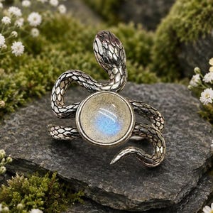 Peut inclure: Une bague argentée en forme de serpent enroulé avec une pierre cabochon ronde et irisée au centre. Le serpent présente des textures d'écailles détaillées sur tout le corps et la tête, la pierre affichant un reflet de lumière bleue.