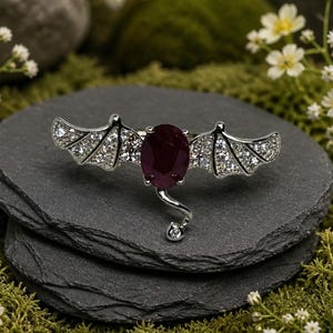 May include: A silver-toned ring featuring a central oval-cut deep red gemstone flanked by two bat-wing shaped accents encrusted with small clear stones. A thin silver tail extends downward from the center, ending in a single clear stone.