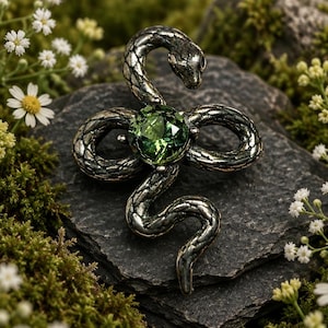 May include: A silver-toned metal brooch shaped like a coiled snake with a textured, scale-like surface. The snake is positioned in a loop, featuring a single, round, faceted olive-green gemstone set securely in the centre of its body.