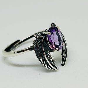 May include: A silver ring featuring a pear-shaped, purple gemstone. The ring's design includes two detailed feather accents on either side of the gemstone. The ring is set against a white background, highlighting its intricate details.