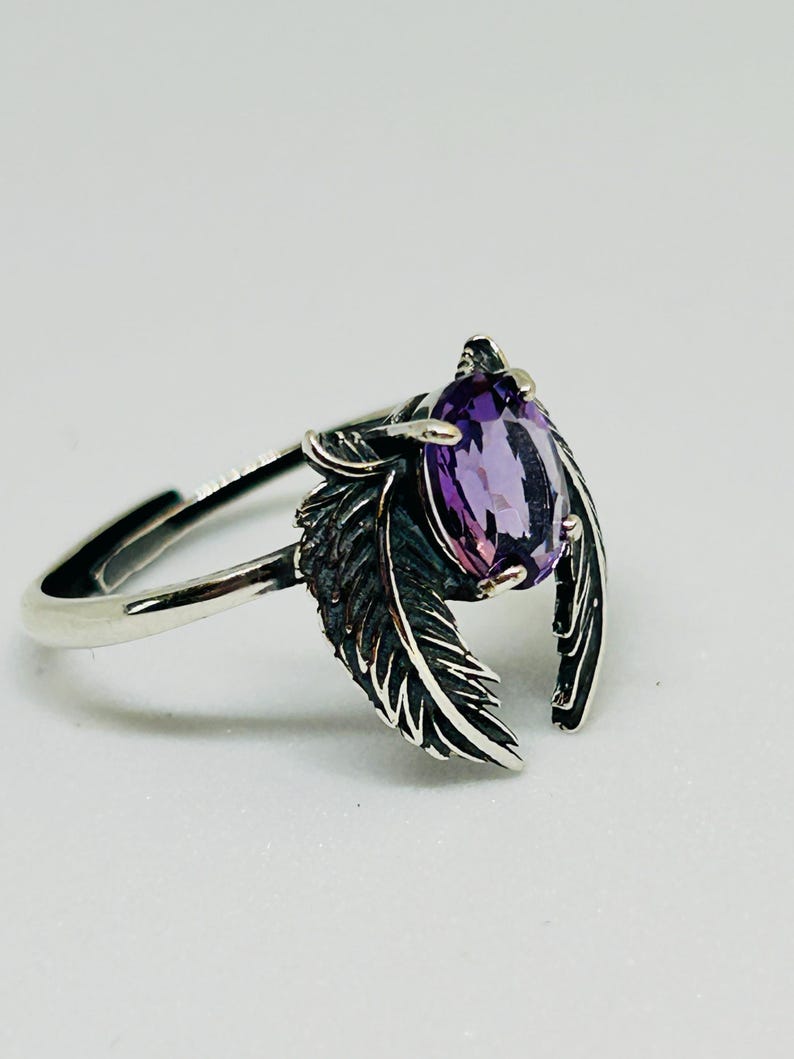 May include: A silver ring featuring a pear-shaped, purple gemstone. The ring's design includes two detailed feather accents on either side of the gemstone. The ring is set against a white background, highlighting its intricate details.