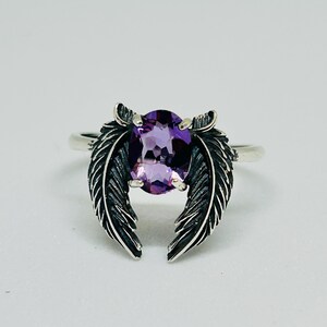 May include: A silver ring featuring a large, oval-cut purple gemstone. The ring's design includes two detailed feather accents on either side of the gemstone, creating a unique and elegant look. The ring is set against a plain white background.