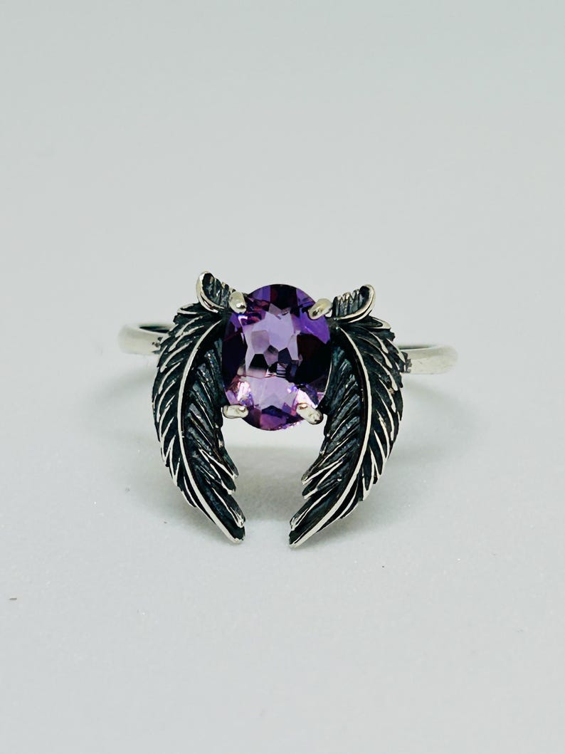 May include: A silver ring featuring a large, oval-cut purple gemstone. The ring's design includes two detailed feather accents on either side of the gemstone, creating a unique and elegant look. The ring is set against a plain white background.