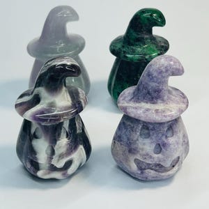 May include: Four carved stone pumpkin figurines wearing witch hats. The pumpkins are in shades of green, purple, and grey. Each pumpkin has a carved face. The figurines are set against a white background.