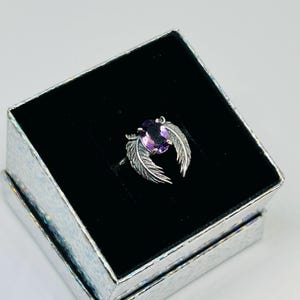 May include: A silver ring with a purple oval gemstone, flanked by feather designs, rests in a black velvet-lined box. The box is silver with a textured, iridescent finish, creating a striking presentation for the jewelry.