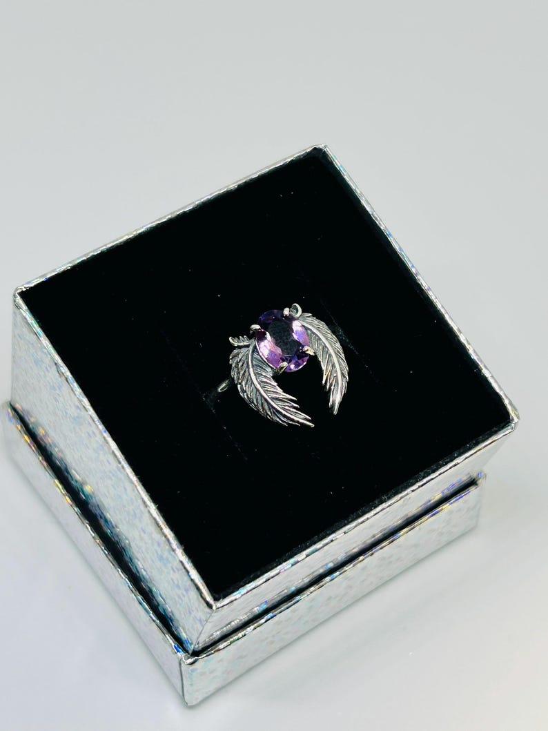 May include: A silver ring with a purple oval gemstone, flanked by feather designs, rests in a black velvet-lined box. The box is silver with a textured, iridescent finish, creating a striking presentation for the jewelry.