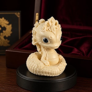 May include: Ivory dragon figurine with blue eyes, seated on a circular base. The dragon has detailed scales and floral accents. The figurine is displayed on a black stand, with a dark wooden box and red velvet interior in the background.