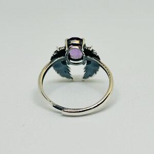 May include: A silver-toned ring featuring an oval, light purple gemstone. The ring has a decorative design on either side of the gemstone, resembling stylized wings or leaves. The band is adjustable.