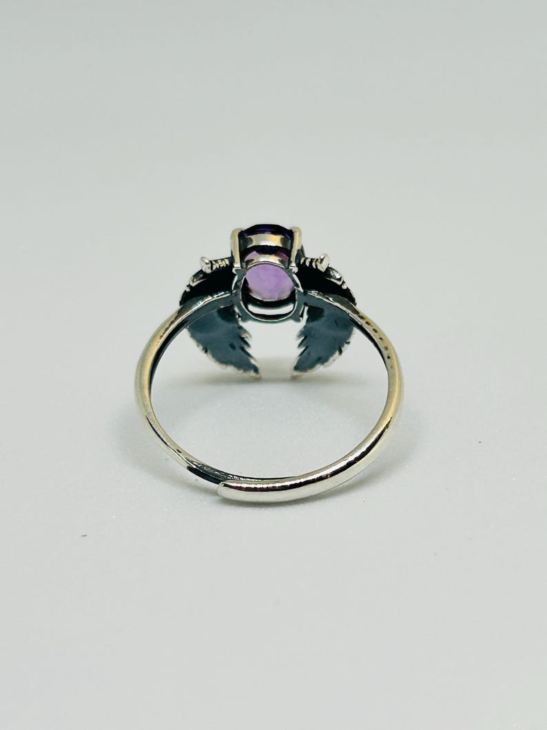 May include: A silver-toned ring featuring an oval, light purple gemstone. The ring has a decorative design on either side of the gemstone, resembling stylized wings or leaves. The band is adjustable.