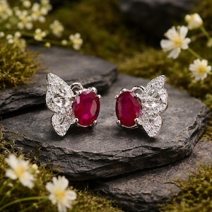May include: A pair of butterfly-shaped earrings with ruby-red oval gemstones and sparkling white accents. The earrings are set on a dark gray stone surface, surrounded by white flowers and green moss.