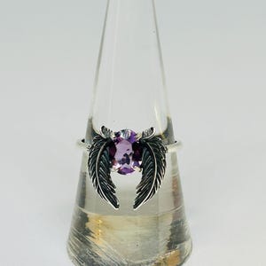 May include: A silver ring featuring a purple oval gemstone, flanked by two detailed feather designs. The ring is displayed on a clear, conical stand against a white background. The ring's design is intricate and elegant.